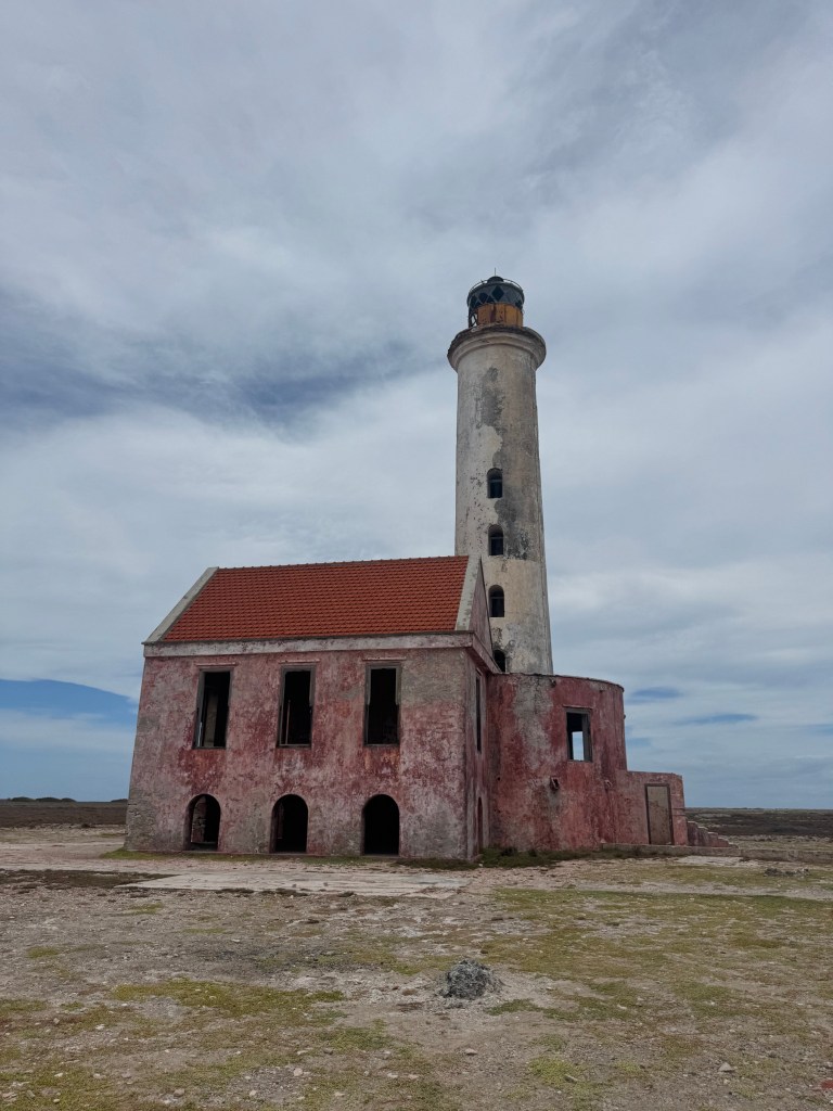 A weathered lighthouse with a red-tiled roof, standing tall beside an ancient building, amidst a cloudy sky and barren landscape.