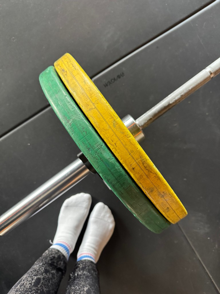 Close-up view of a weightlifting barbell with green and yellow plates, with feet in white socks visible in the foreground.