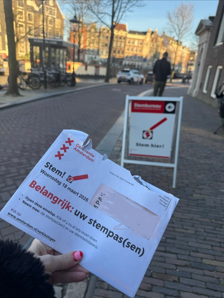 A person holding an election envelope in Amsterdam, with buildings and a street visible in the background, and a voting sign nearby.