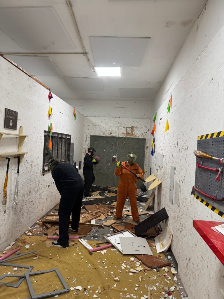 Two individuals in protective gear are breaking objects in a messy room filled with debris. One person is wearing an orange jumpsuit and holding a sledgehammer, while the other is in black attire. Colorful decorations are visible on the walls.