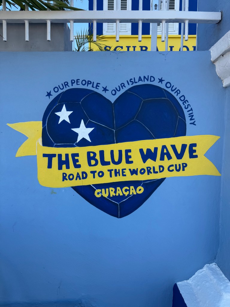 A mural featuring a heart-shaped design with a soccer ball, promoting the phrase 'THE BLUE WAVE ROAD TO THE WORLD CUP CURAÇAO' along with 'OUR PEOPLE * OUR ISLAND * OUR DESTINY'.