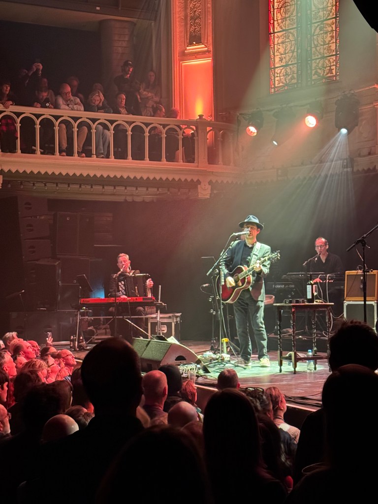 Neil Hannon of the Divine Comedy performs on stage with an acoustic guitar, surrounded by musicians and a captivated audience in a well-lit venue.