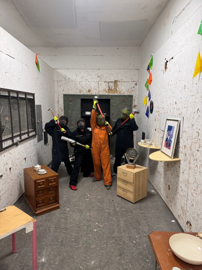 Four people in protective gear holding various tools in a cluttered indoor space, featuring damaged walls and furniture.