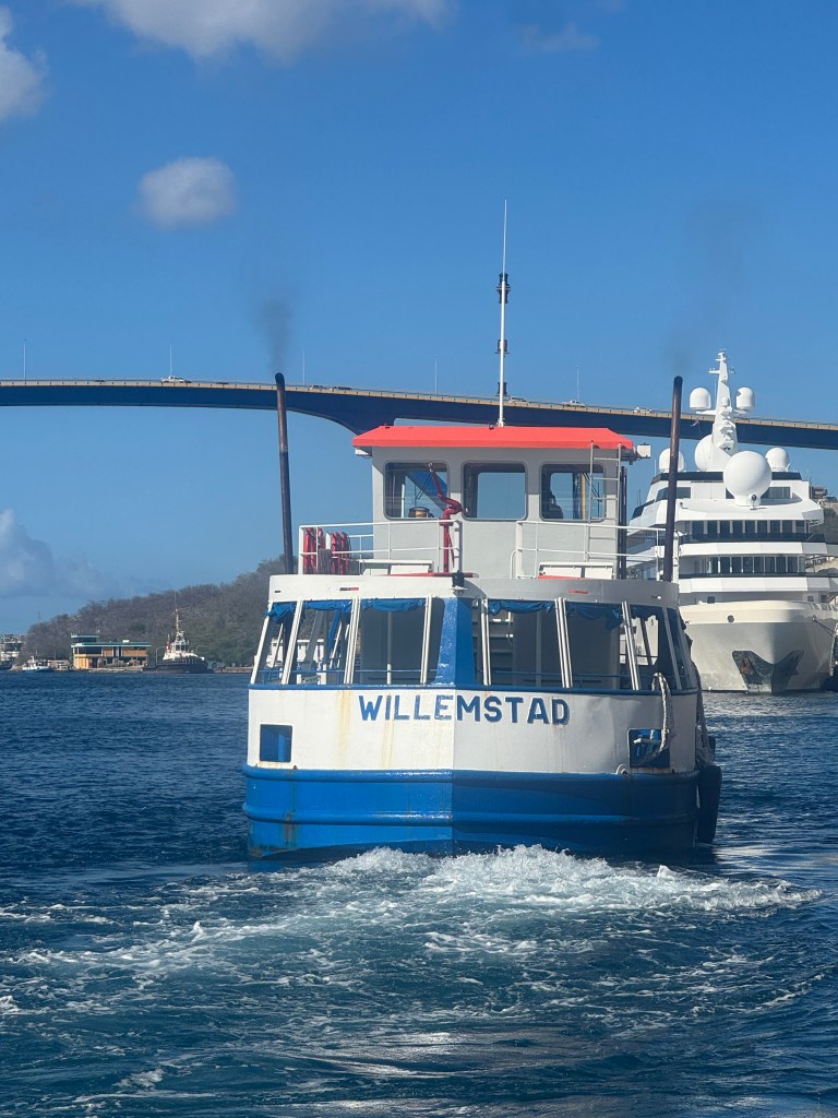 A small boat named 'Willemstad' cruising through blue water, with a large ship and a bridge in the background.