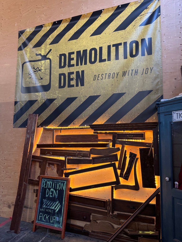 A large banner reading 'DEMOLITION DEN' with the tagline 'DESTROY WITH JOY', set against a backdrop of wooden panels. Below, a chalkboard sign directs visitors to 'WAIT HERE' and 'PICK UP', surrounded by illuminated wooden pieces.