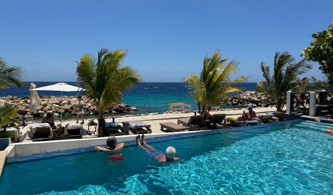 View of a tropical resort pool with two people relaxing in the water, surrounded by palm trees and a beach with lounge chairs in the background.
