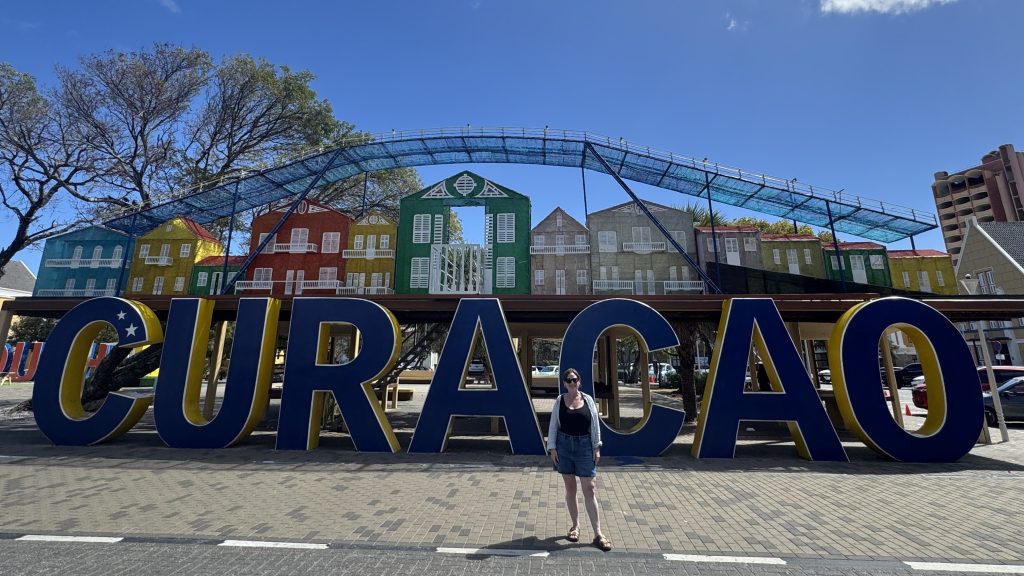 Colorful landmark sign reading 'CURACAO' with a woman standing in front, surrounded by vibrant houses and a clear blue sky.